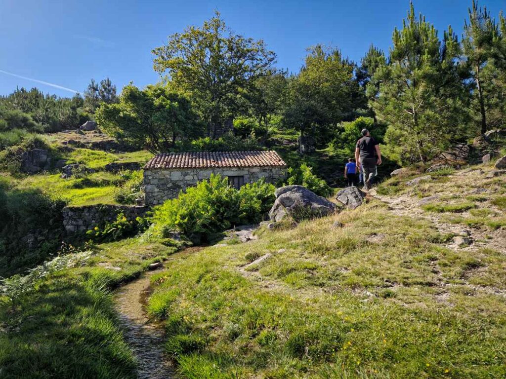 Caminho inserido na natureza com uma casa de pedra ao fundo e pessoas a andar