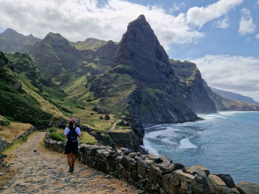 Pessoa a caminhar num trilho de paisagem de montanha verde ao fundo ao pé do mar, em Santo Antão, Cabo Verde.