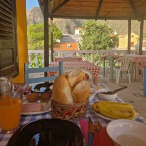 Mesa de pequeno-almoço com sumo, bananas, pão e bolos, num varanda com várias mesas, no alojamento Aldeia Jerome no Paul