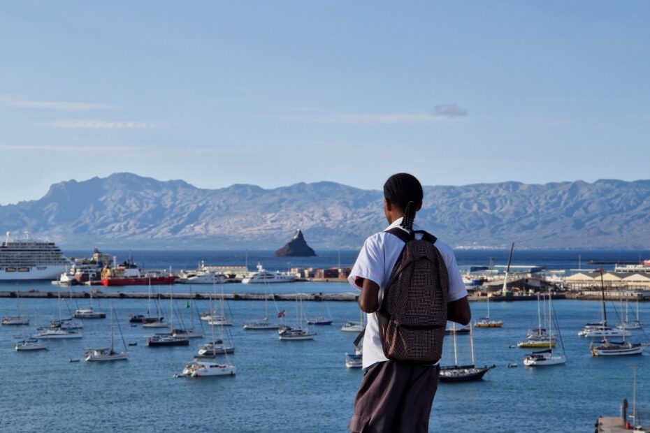 Rapaz de Cabo Verde de camisa branca e mochila às cosas a olhar o mar azul com muitos barcos e montanhas ao fundo