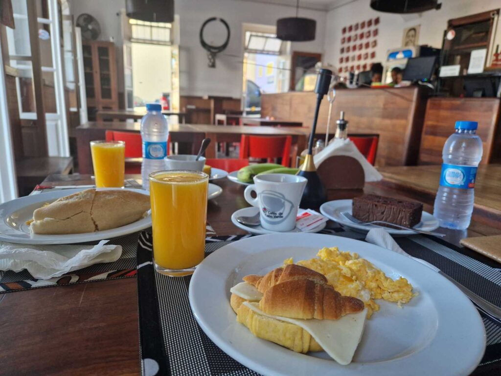 Mesa de pequeno-almoço do alojamento em Mindelo com croissant, ovos mexidos, pão, sumos, café, bolo e água