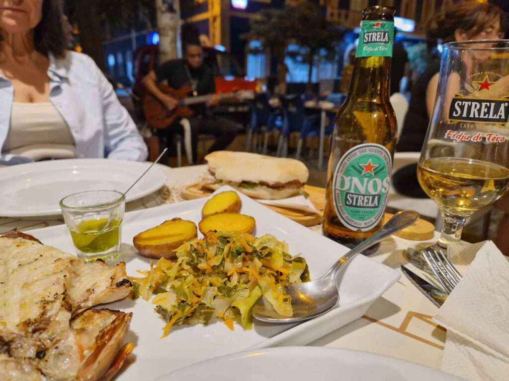 Prato de peixe com batatas e legumes, ao lado de uma cerveja Strela, e ao fundo uma pessoa a tocar guitarra, no Casa Café Mindelo
