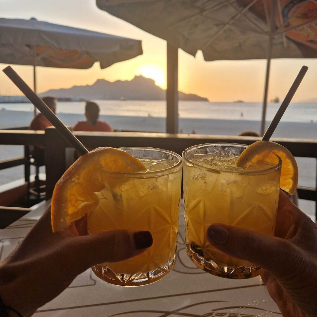 Dois copos com bebida laranja numa esplanada com o por do sol o fundo, na Praia da Laginha em Mindelo, Cabo Verde