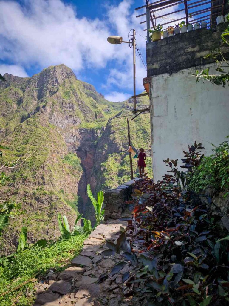 Casa em penhasco da aldeia da Vinha em Santo Antão com uma pessoa em cima de um muro e montanhas verdes ao fundo