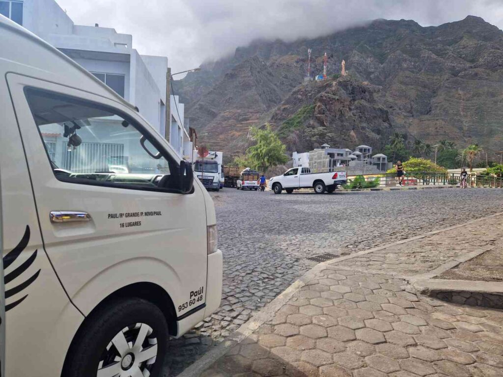 Carrinha de transporte coletivo em largo amplo do Paúl, em Santo Antão, com uma montanha e nuvens ao fundo