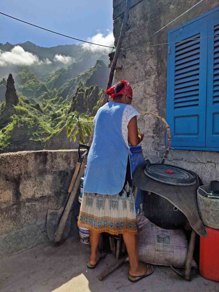 Senhora com avental azul e lenço vermelho à cabeça de costas em terraço com materiais de construção e montanhas verdes ao fundo