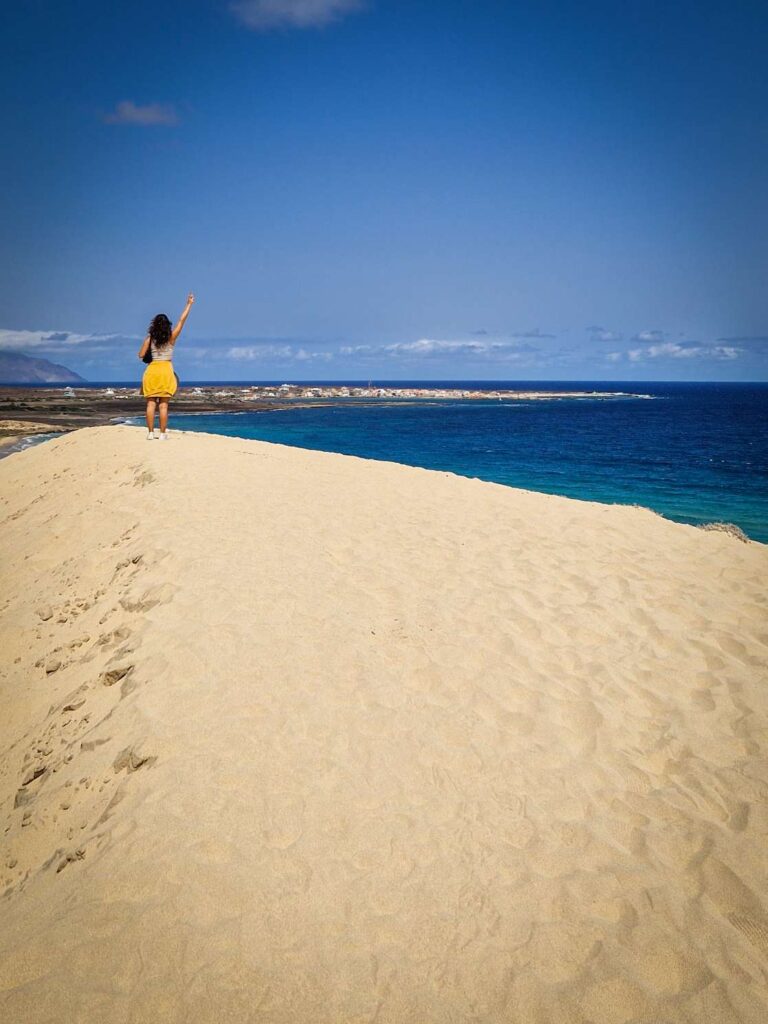 Pessoa com braço no ar ao fundo de uma duna de areia branca e um mar azul na paisagem