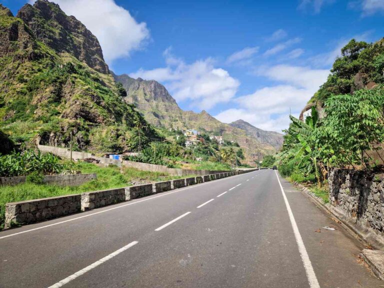 Estrada alcatroada entre montanhas com algumas casas coloridas ao fundo debaixo de céu azul salpicado de nuvens, Santo Antão, Cabo Verde