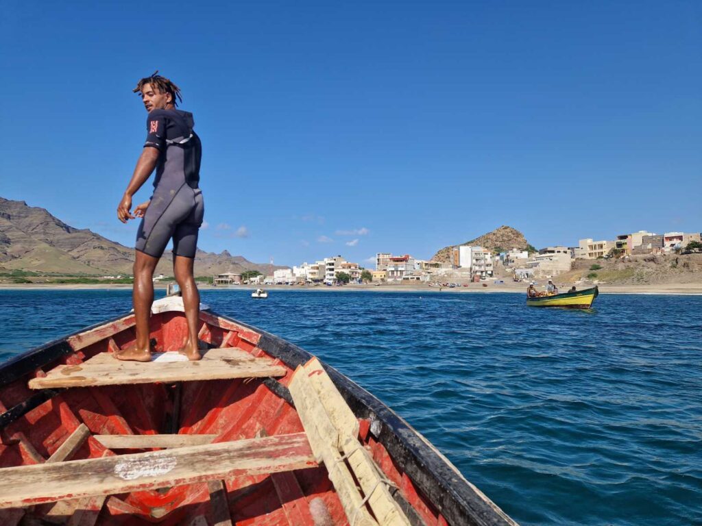 Jovem em cima de barco de madeira vermelho no mar azul, ao fundo praia e edifícios