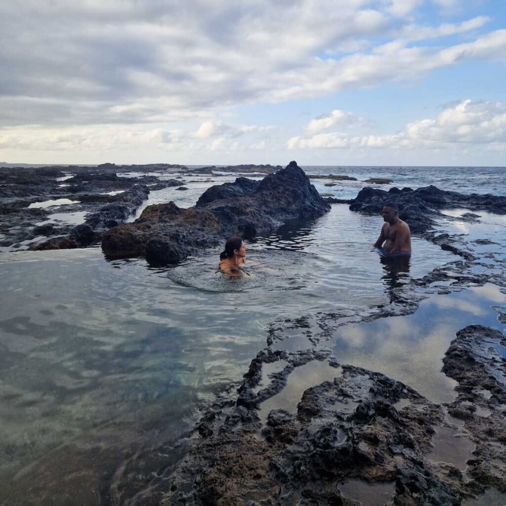 Duas pessoas a conversar sentadas na água entre as rochas das Piscinas Naturais de Sinagoga em Santo