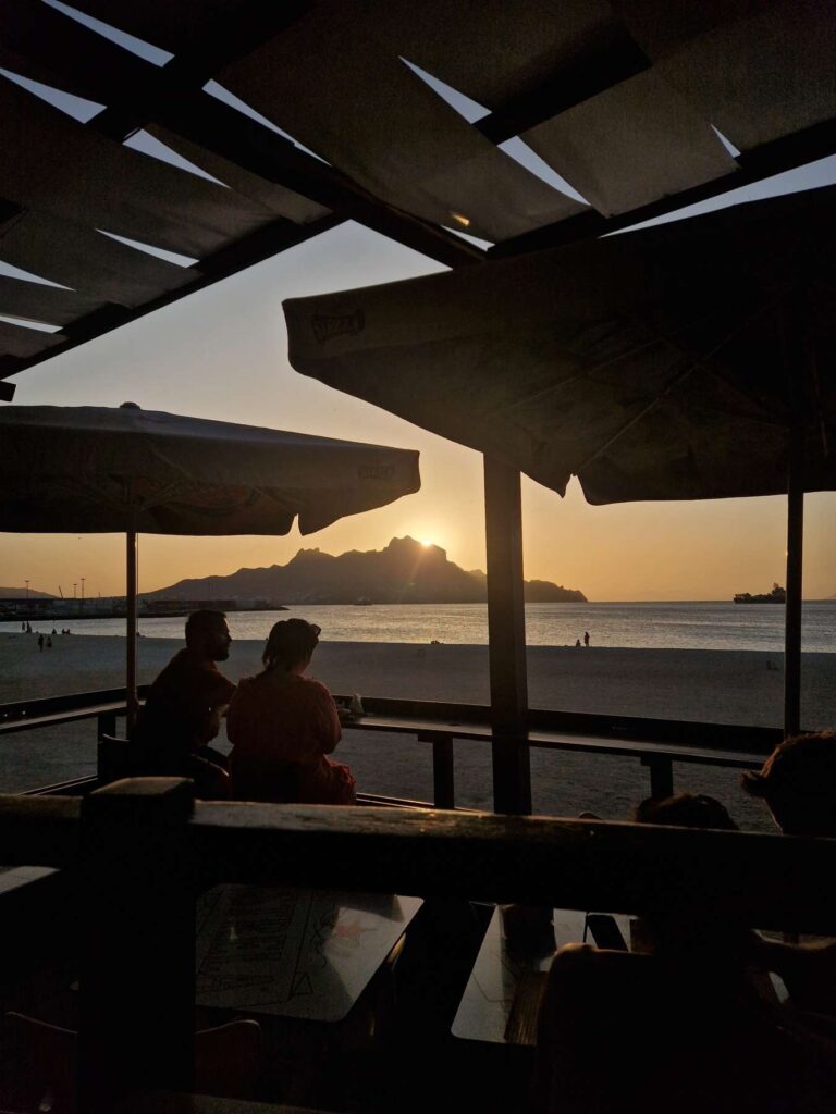 Vista do por do sol sobre o Monte Cara numa esplanada com pessoas na Praia da Laginha, no Mindelo