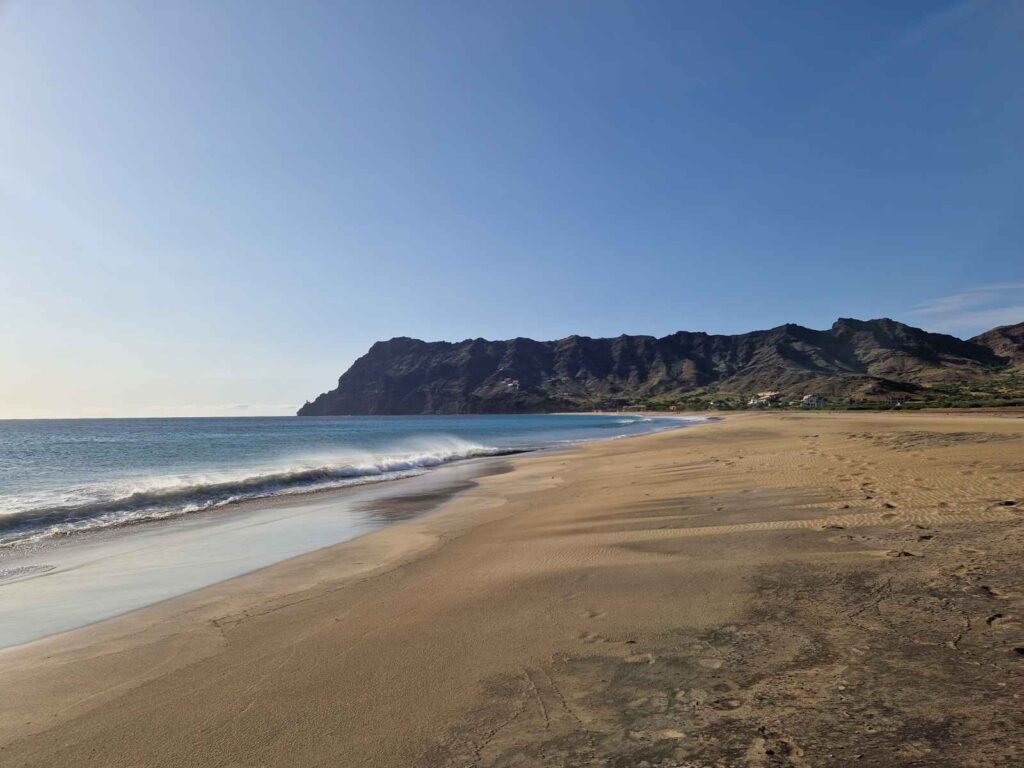 Praia extensa de areia amarela e rebentação das ondas fortes do mar, uma montanha castanha ao fundo