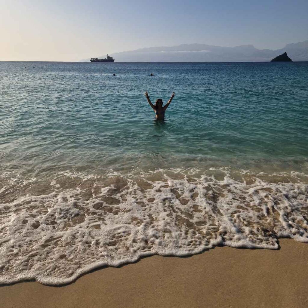 Pessoa com braços levantados no mar azul junto á rebentação branca das ondas sobre a areia, na Praia da Laginha, Mindelo, Cabo Verde