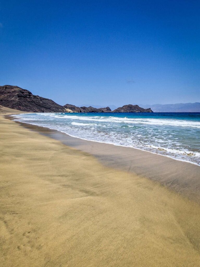 Praia de areia amarela e mar azul com algumas ondas e rochas ao fundo