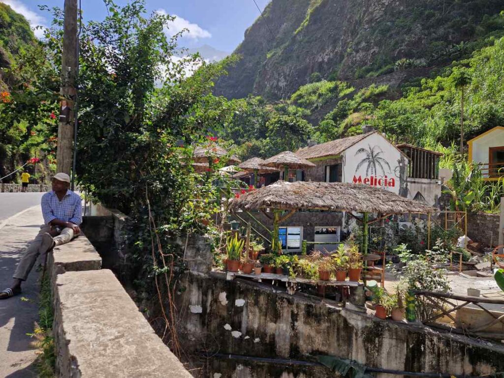 Restaurante Melícia com telhados de palha ao pé de uma estrada com um senhor de chapéu sentado num muro