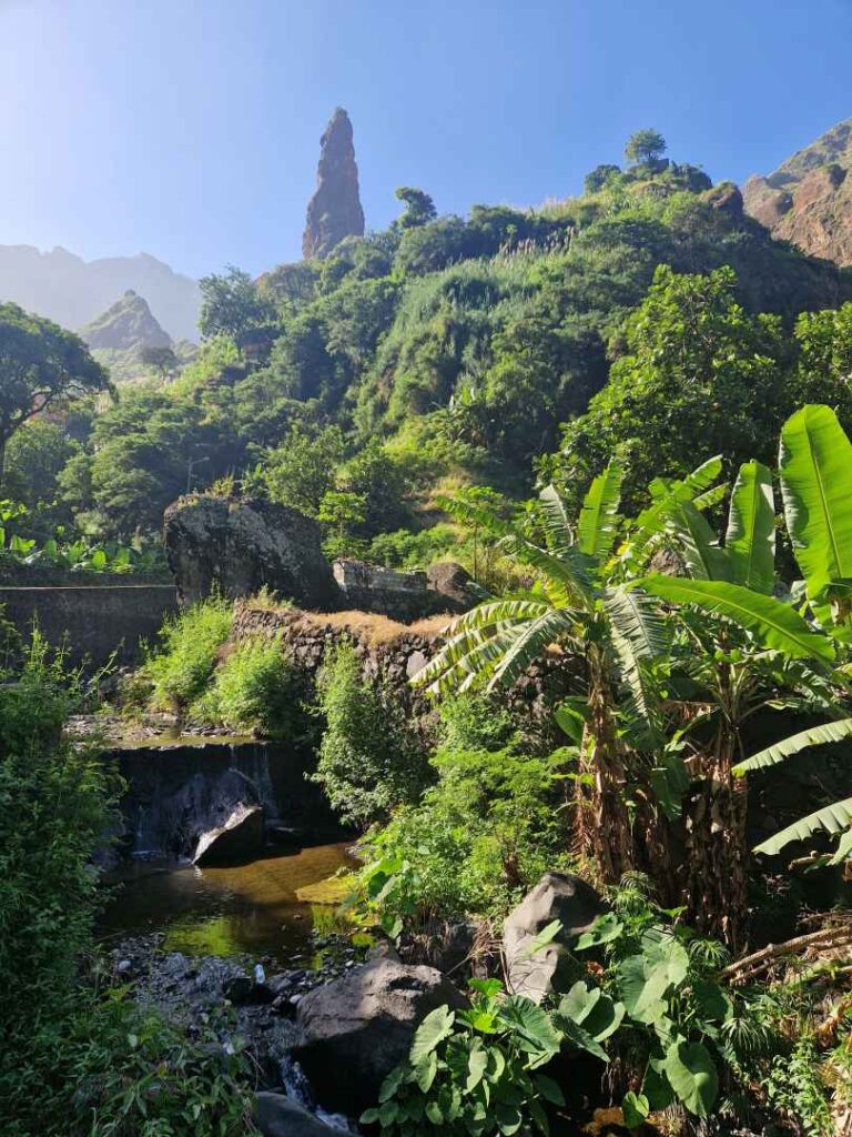 Paisagem verde de Santo Antão com um ribeiro em baixo à esquerda, o Lombo do Pico ao fundo em cima, pela frente de um céu azul