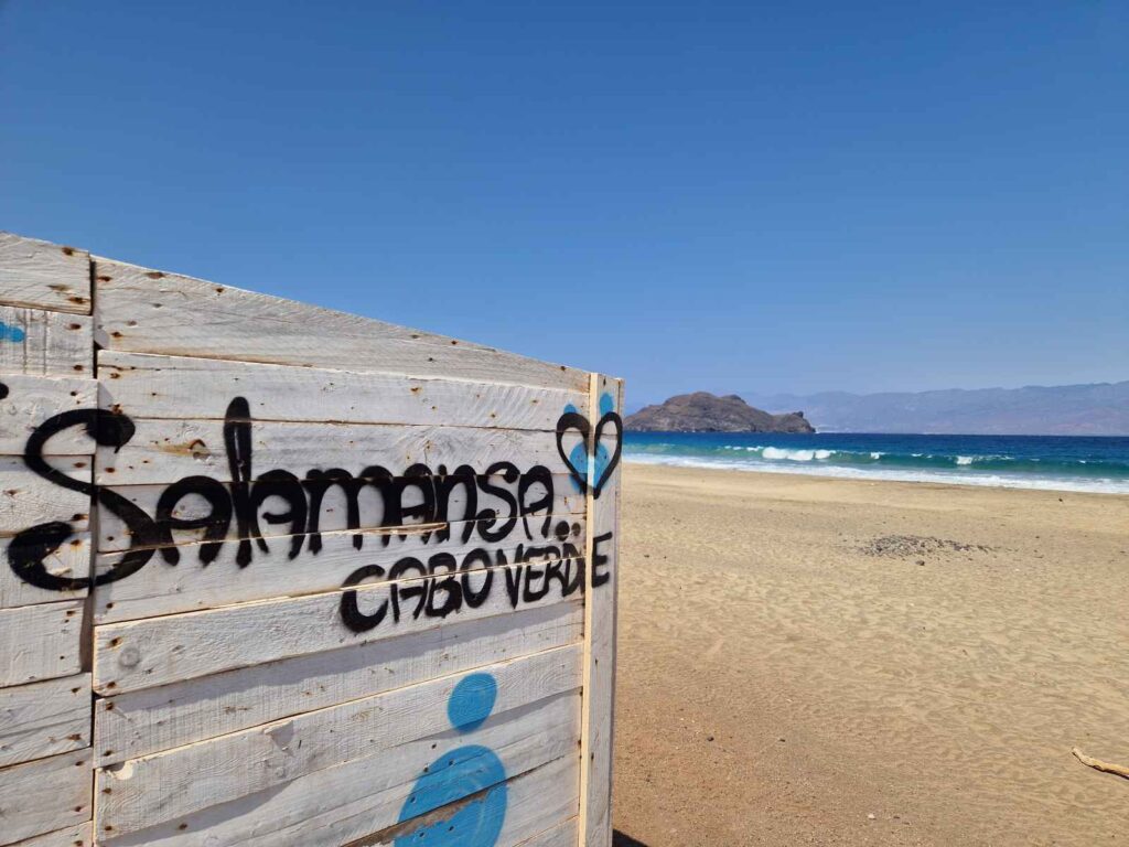 Parede de madeira com grafitti a dizer Salamansa Cabo Verde junto à praia de areia amarela e mar azul