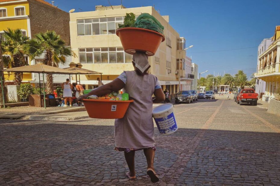 Senhora a caminhar com uma cesta de legumes à cabeça, outra num braço e um balde no outro, numa rua do Mindelo, em São Vicente, Cabo Verde