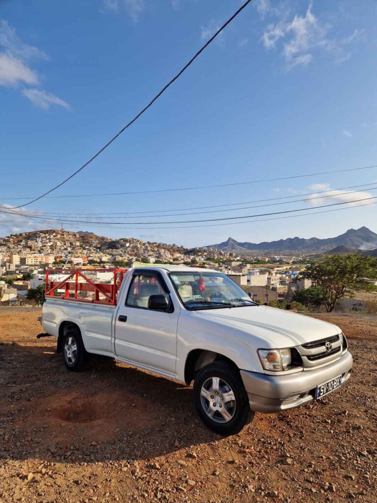 Carro Toyota branco com parte traseira vermelha numa estrada em pedra, a cidade de Mindelo e as montanhas de São Vicente ao fundo